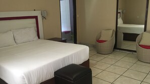 Een hotelkamer met een bed, een nachtkastje, een zwart poufje, twee rieten stoelen met rode kussens en een grote spiegel.