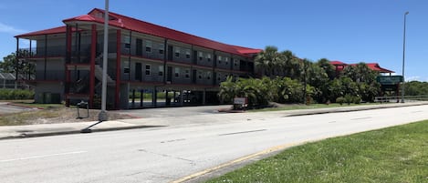 A multi-story hotel with a red roof and balconies, located next to a road with a grassy area in front.