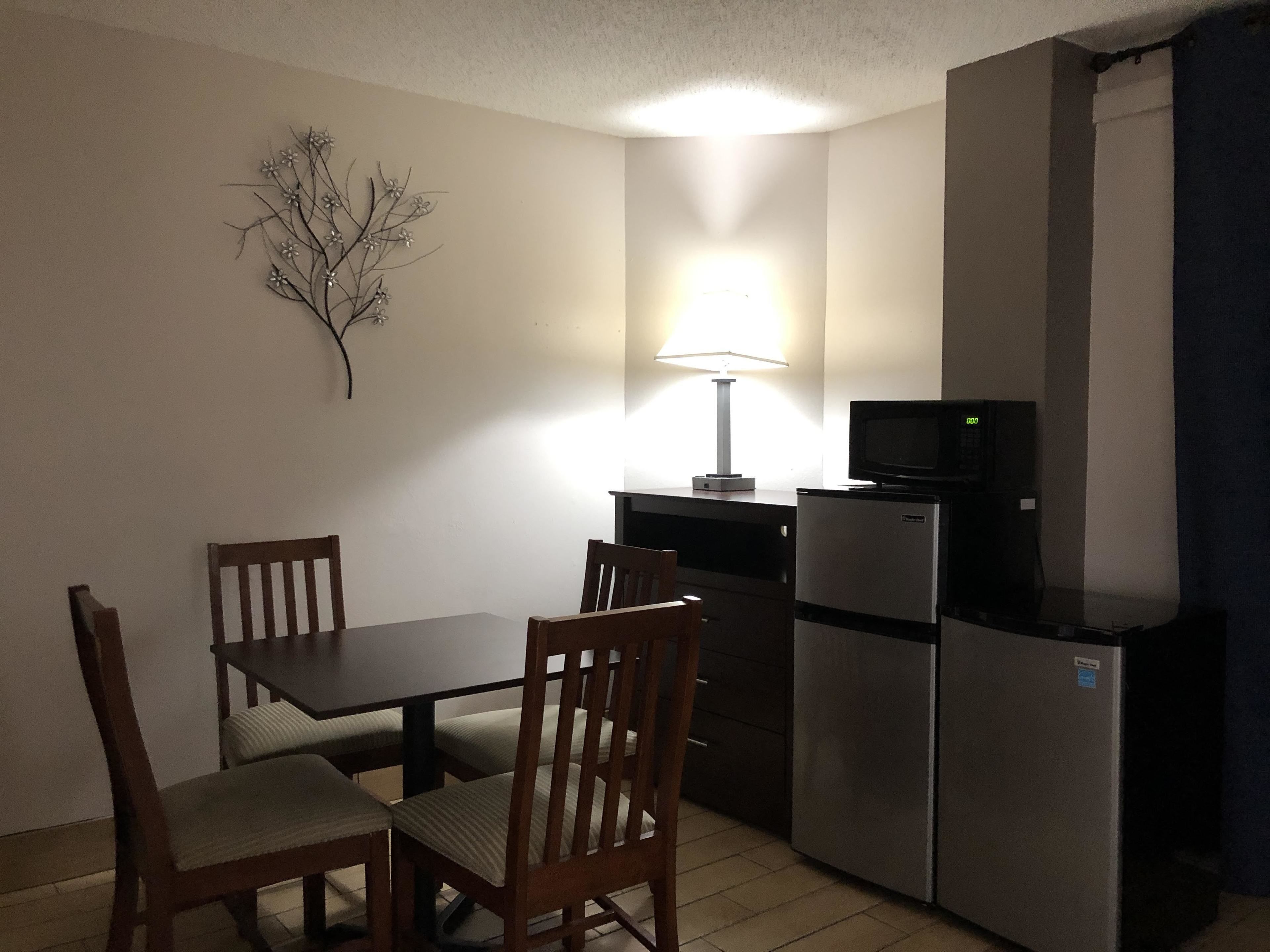 A compact kitchen with a dining table and chairs, a refrigerator, a microwave, and a wall-mounted lamp.
