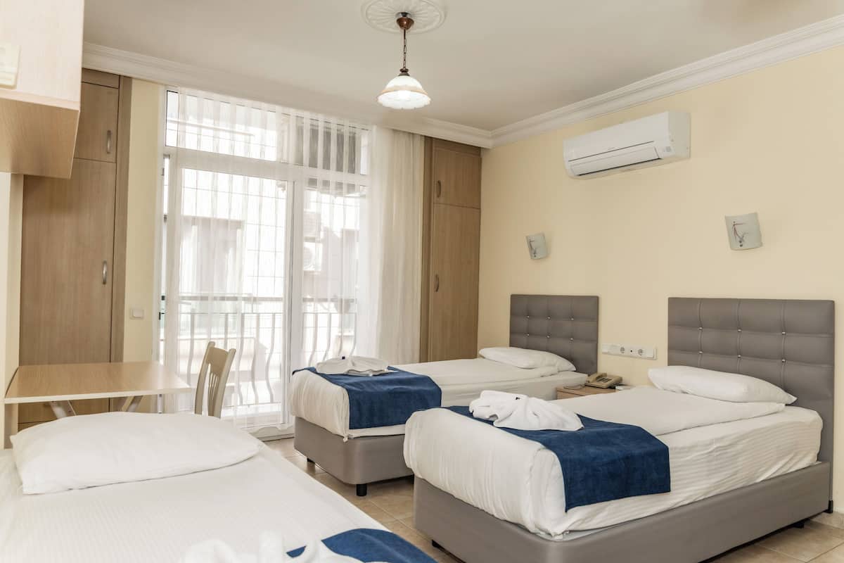 Quarto de hotel com duas camas, uma escrivaninha e uma cadeira. Há uma janela com cortinas, um ar condicionado na parede e um relógio.