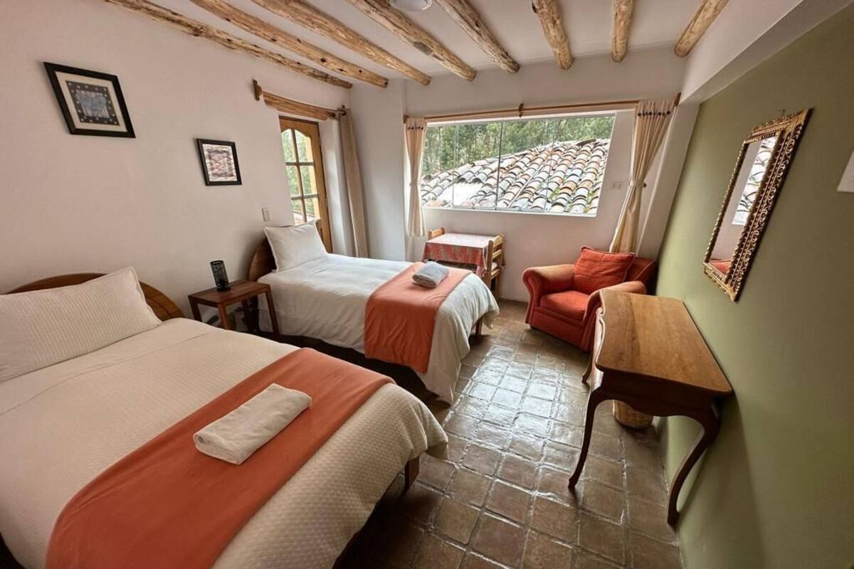 Ein Zimmer mit zwei Betten, einem Holztisch, einem Stuhl und einem Fenster mit Blick auf ein Ziegeldach.