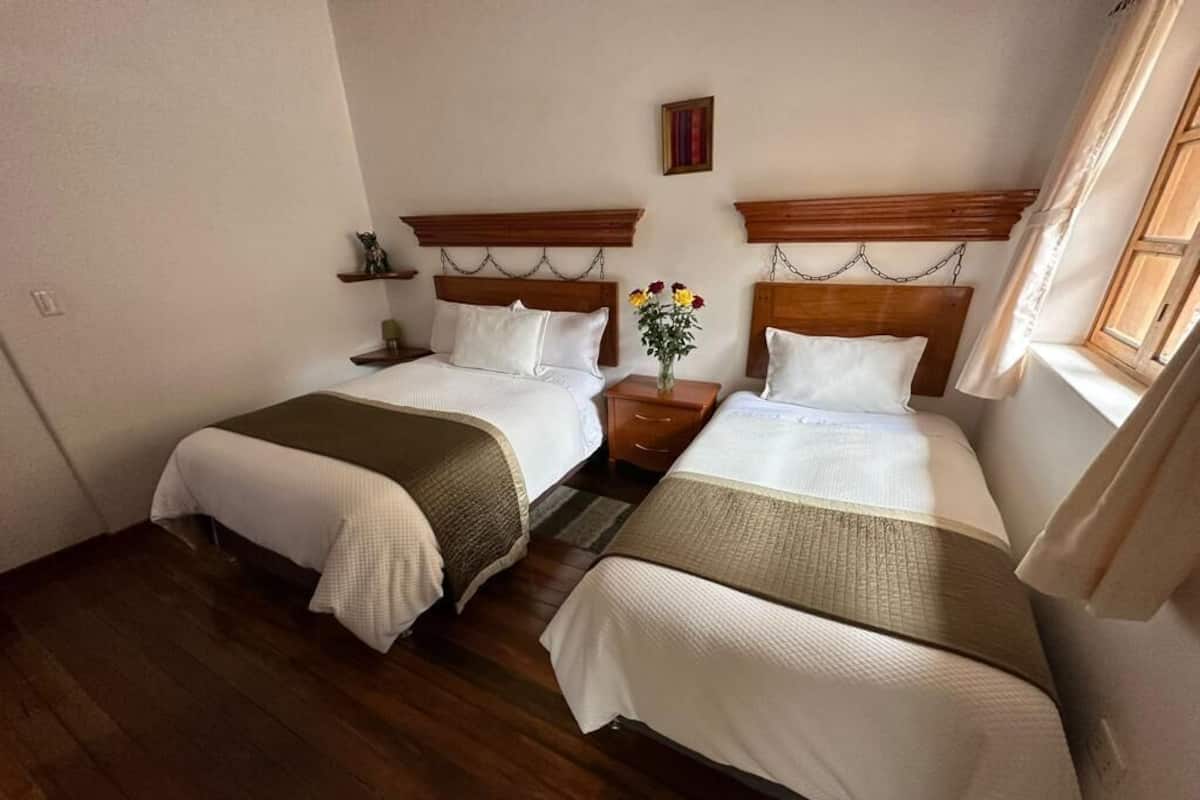 Ein Zimmer mit zwei Betten, Holz-Kopfteilen, einem kleinen Tisch mit Blumenvase und einem Vorhangfenster.