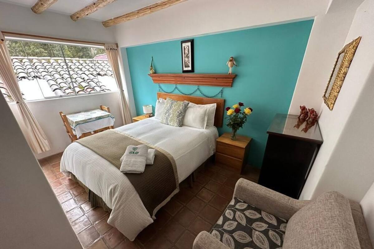 Ein Schlafzimmer mit Bett, Nachttischen, einem Sessel, einem Fenster mit Vorhängen und einem Wandregal mit Dekoration.