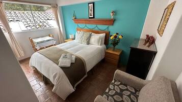 A bedroom with a bed, bedside tables, a chair, a window with curtains, and a wall-mounted shelf with decorative items.