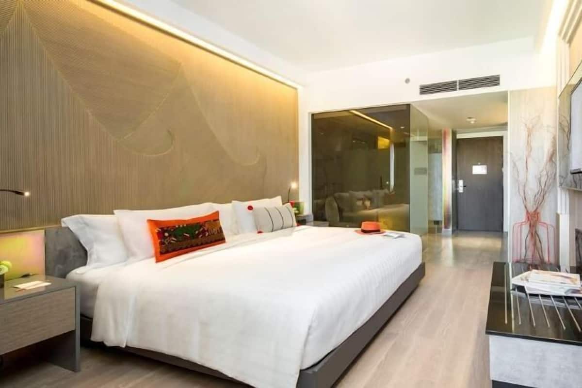 Une chambre d’hôtel moderne dotée d’un grand lit, de tables de chevet et d’une cloison en verre qui sépare la chambre à coucher du salon.