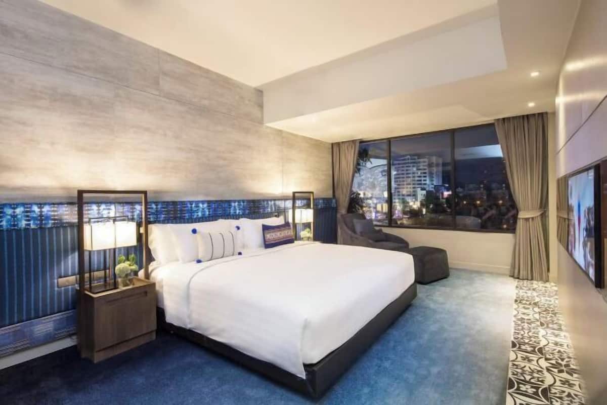 Une chambre d’hôtel moderne avec un grand lit, une table de chevet, une fenêtre donnant sur la ville et une moquette à motifs.