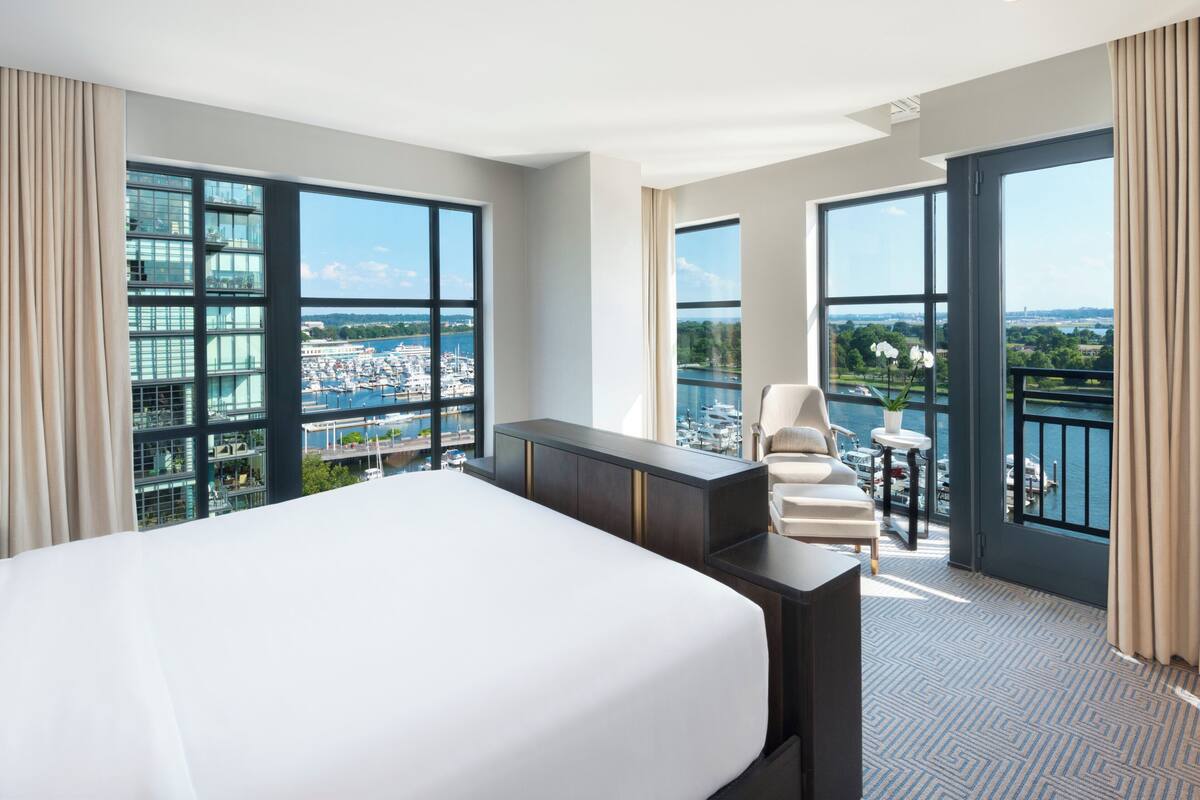 A hotel room with a large bed, a desk, and a chair. It has large windows with a view of a marina and buildings.