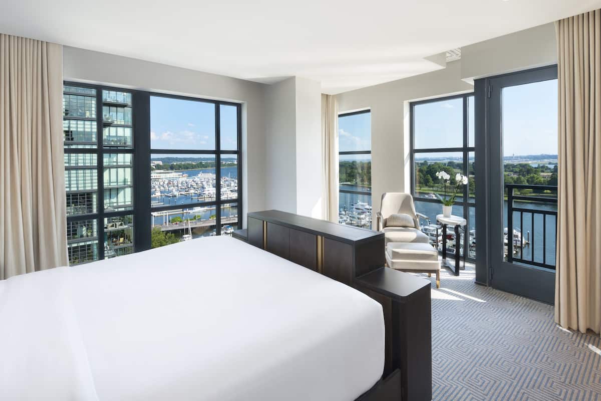 Ein Hotelzimmer mit einem großen Bett, einem Schreibtisch und einem Stuhl. Es hat große Fenster mit Blick auf einen Hafen und Gebäude.