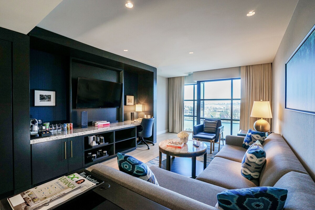 A modern hotel room with a dark wood cabinet, a flat-screen TV, a sofa with patterned cushions, a small round table, and a view of the city through large windows.