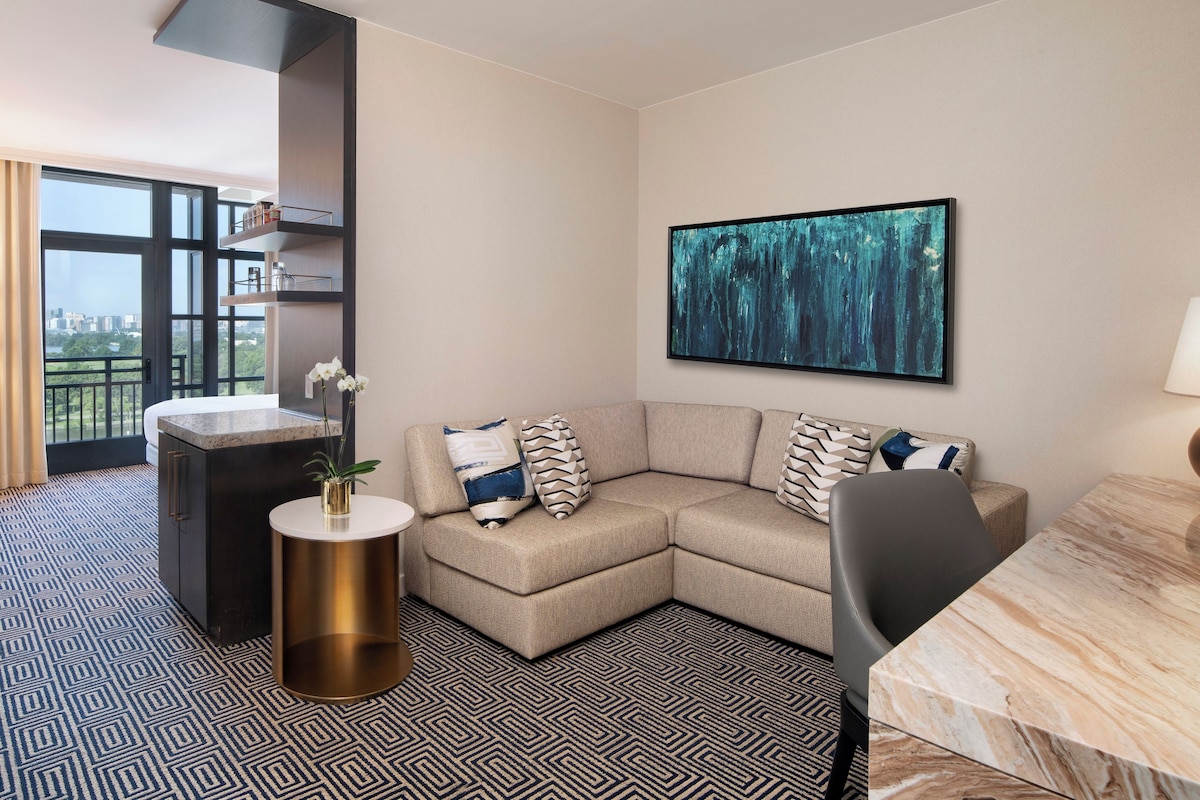 A modern hotel room with a sectional sofa, a round gold side table, a large abstract painting, and a view through a large window.