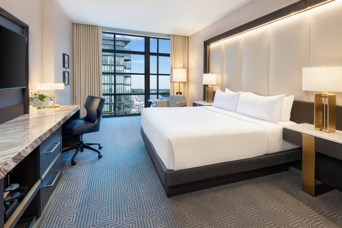 A hotel room with a large bed, a desk with a chair, a TV, and a view of the city.