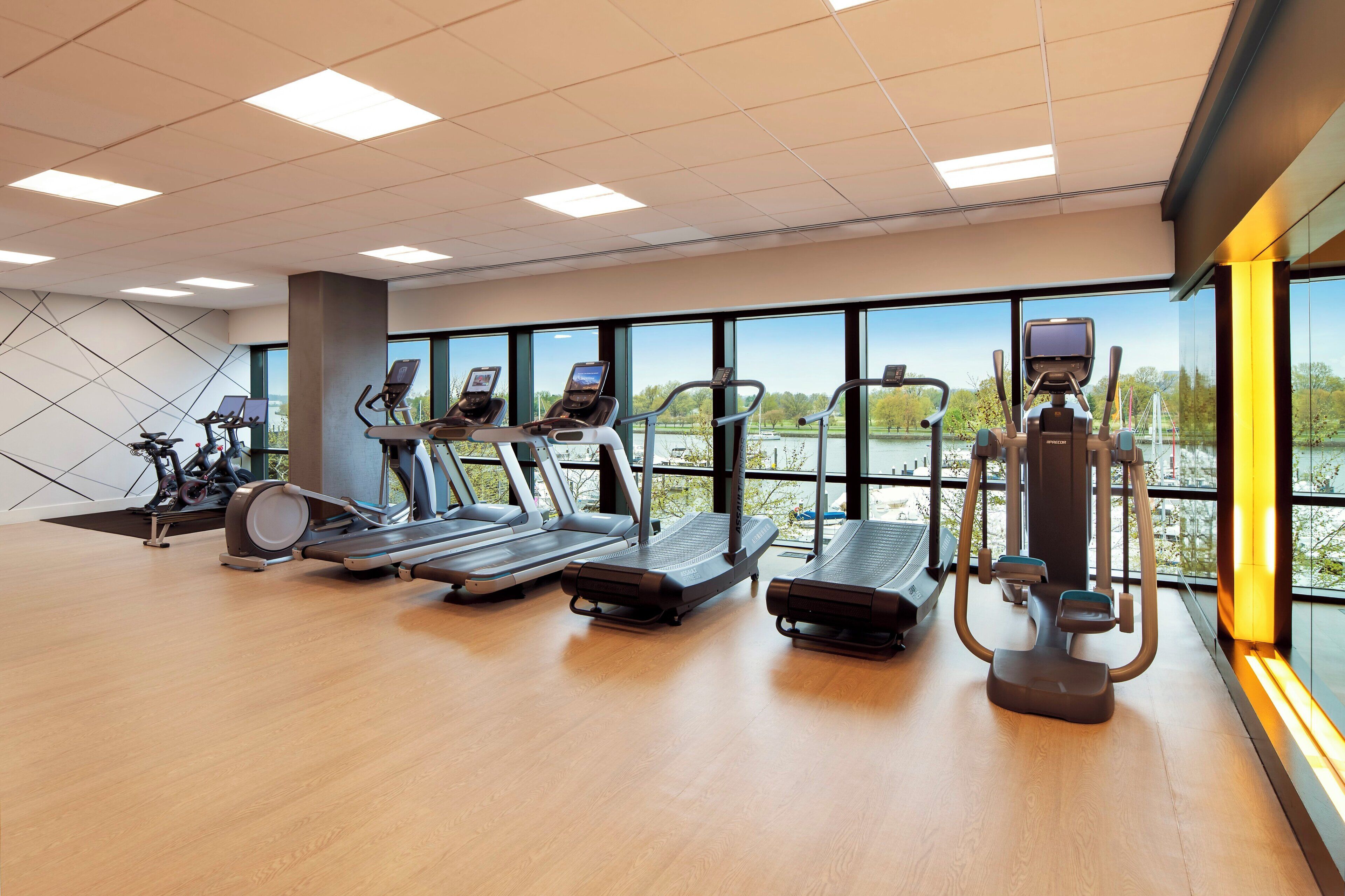 A gym with treadmills, elliptical machines, and a rowing machine, large windows with a view of a park, and a wooden floor.