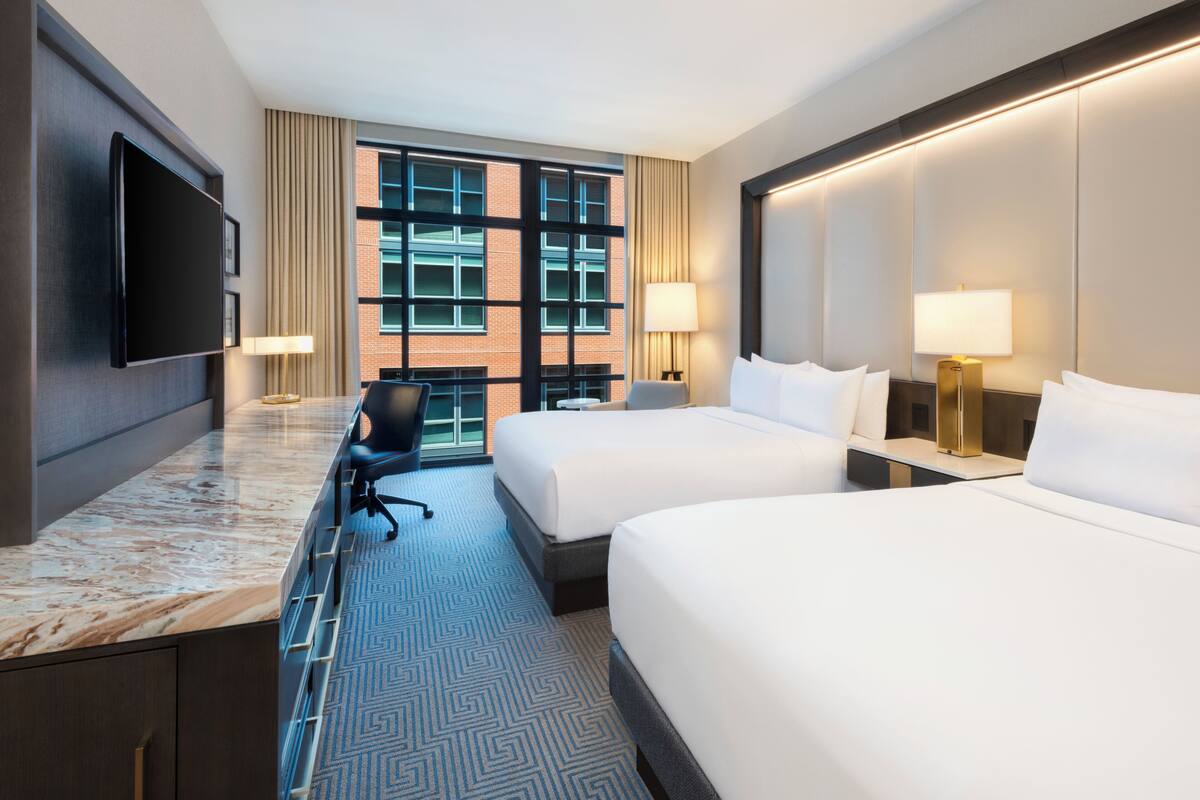 A hotel room with two beds, a desk, a chair, a flat-screen TV, and a large window with a view of a brick building.