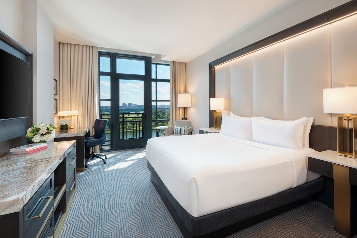A hotel room with a large bed, a desk with a chair, a TV, and a view of the city.
