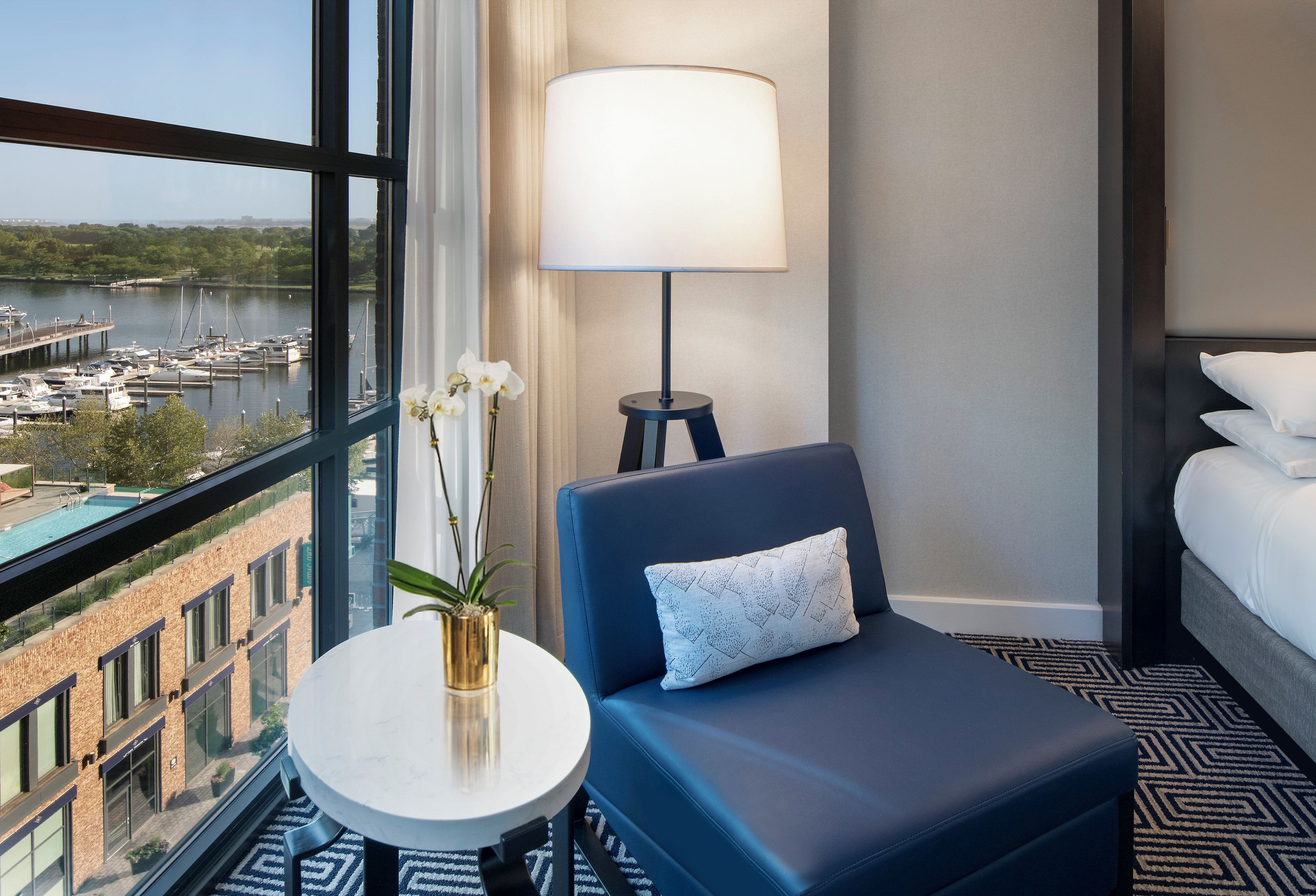Habitación de hotel con vista a un puerto deportivo, una mesa redonda con un florero, un sillón azul con un cojín, una lámpara de pie y una cama con ropa blanca.