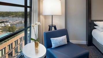 Habitación de hotel con vista a un puerto deportivo, una mesa redonda con un florero, un sillón azul con un cojín, una lámpara de pie y una cama con ropa blanca.