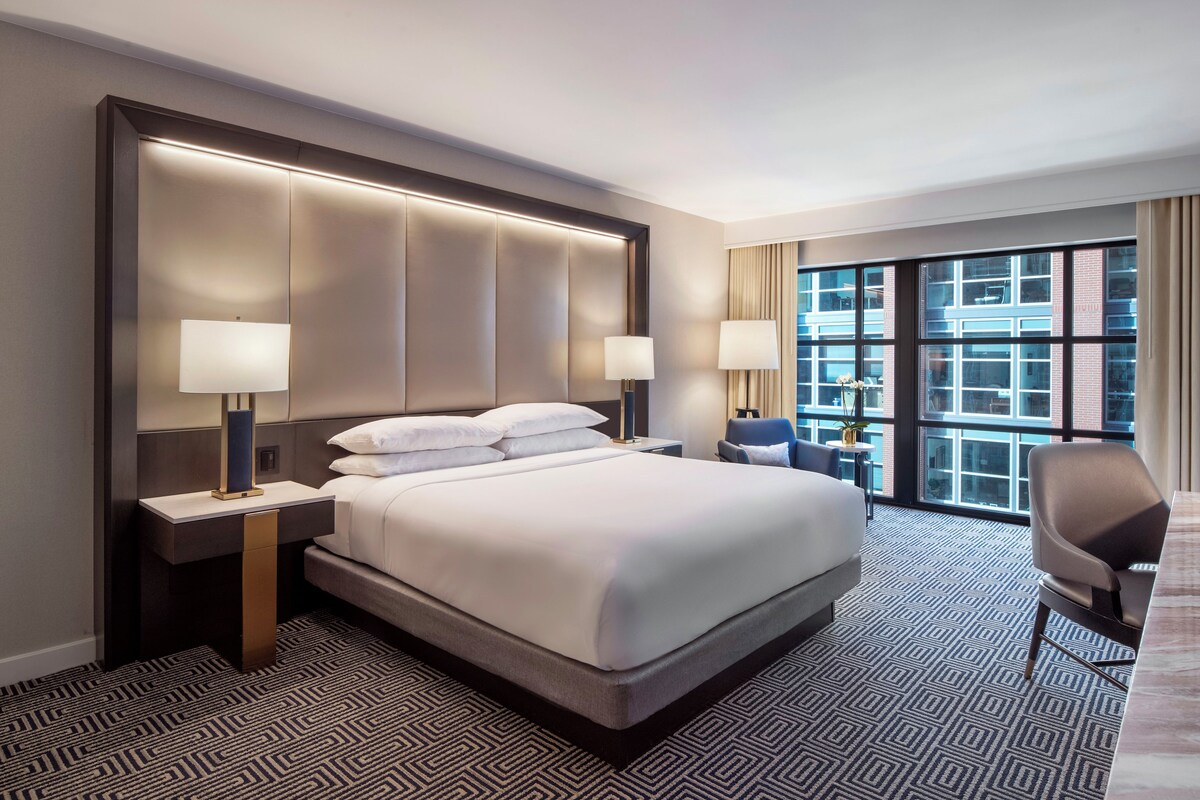 A hotel room with a large bed, two bedside tables, a desk, a chair, and a large window with a view of buildings.