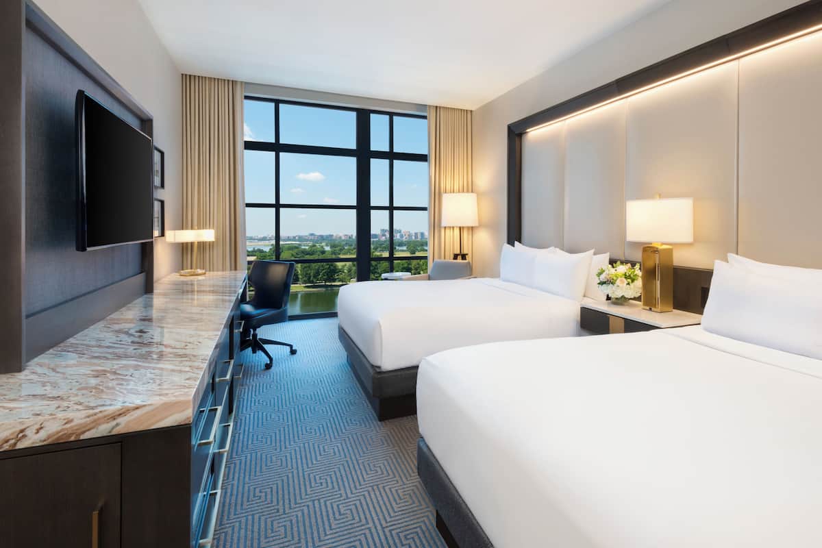 A hotel room with two beds, a desk, a chair, a TV, and a large window with a city view.