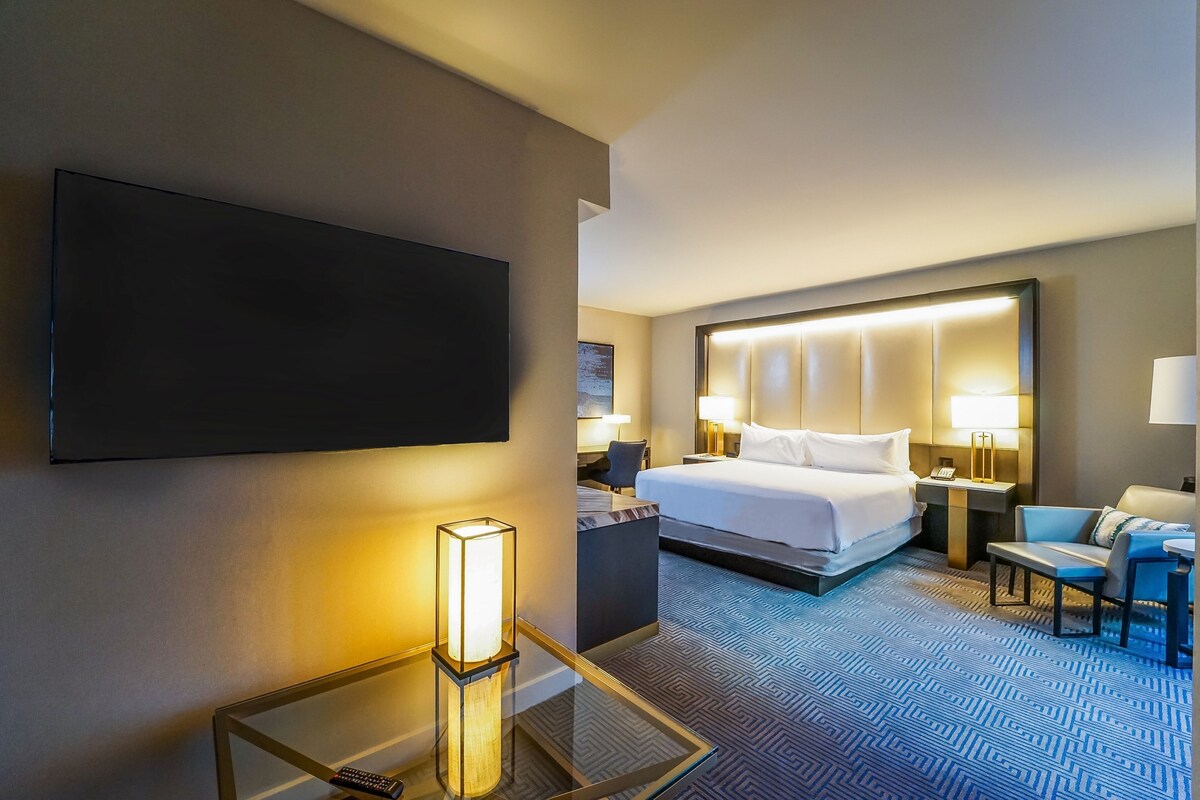 A hotel room with a large bed, a flat-screen TV, a glass coffee table, a desk with a lamp, and a patterned carpet.