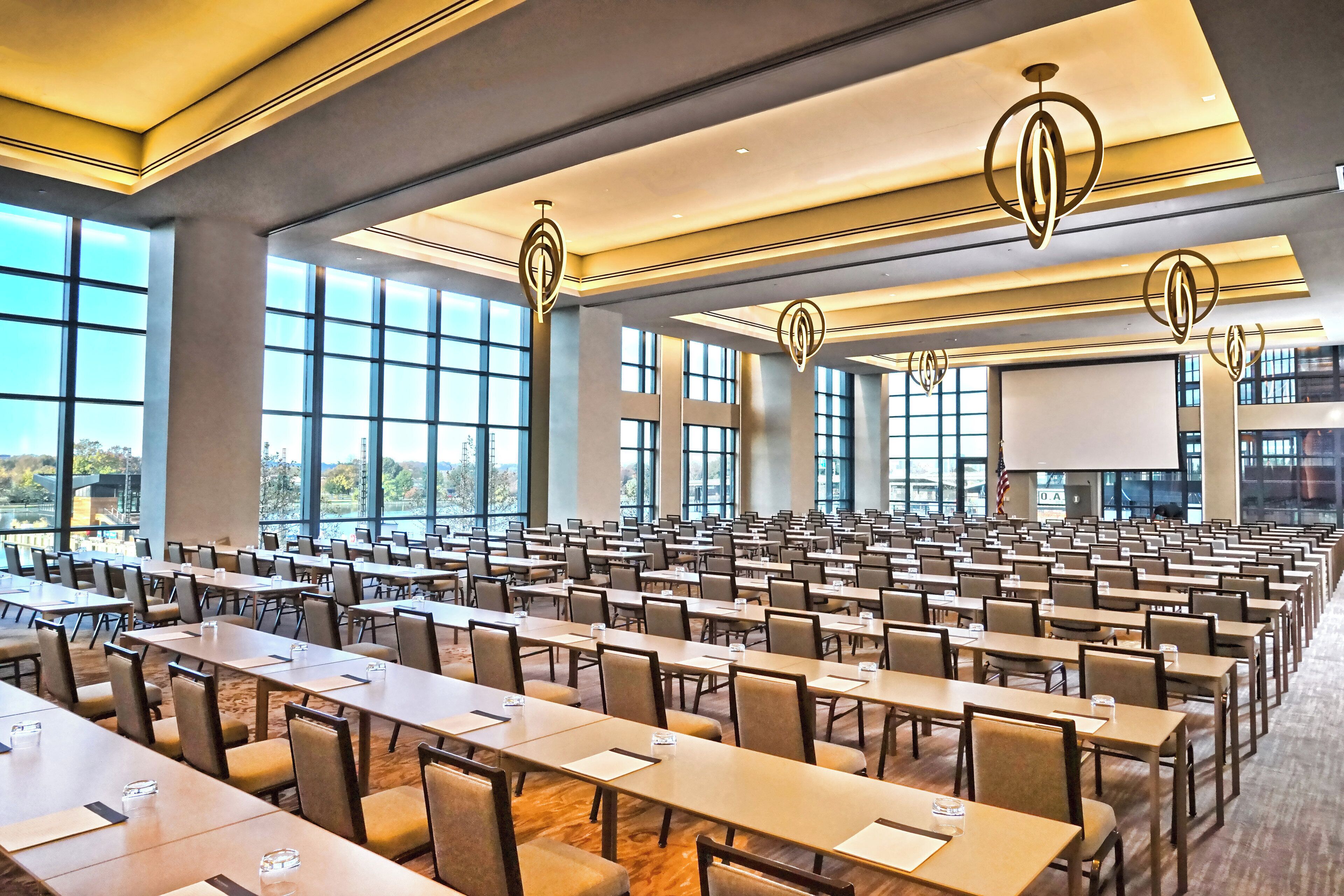 Un amplio salón de conferencias con filas de mesas y sillas, ventanas grandes y luminarias modernas.