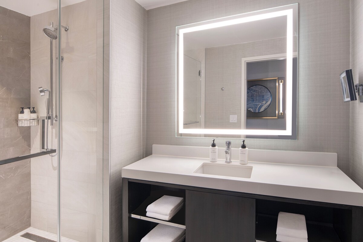 A modern bathroom with a glass shower enclosure, a large mirror with integrated lighting, and a vanity with a sink and towel storage.