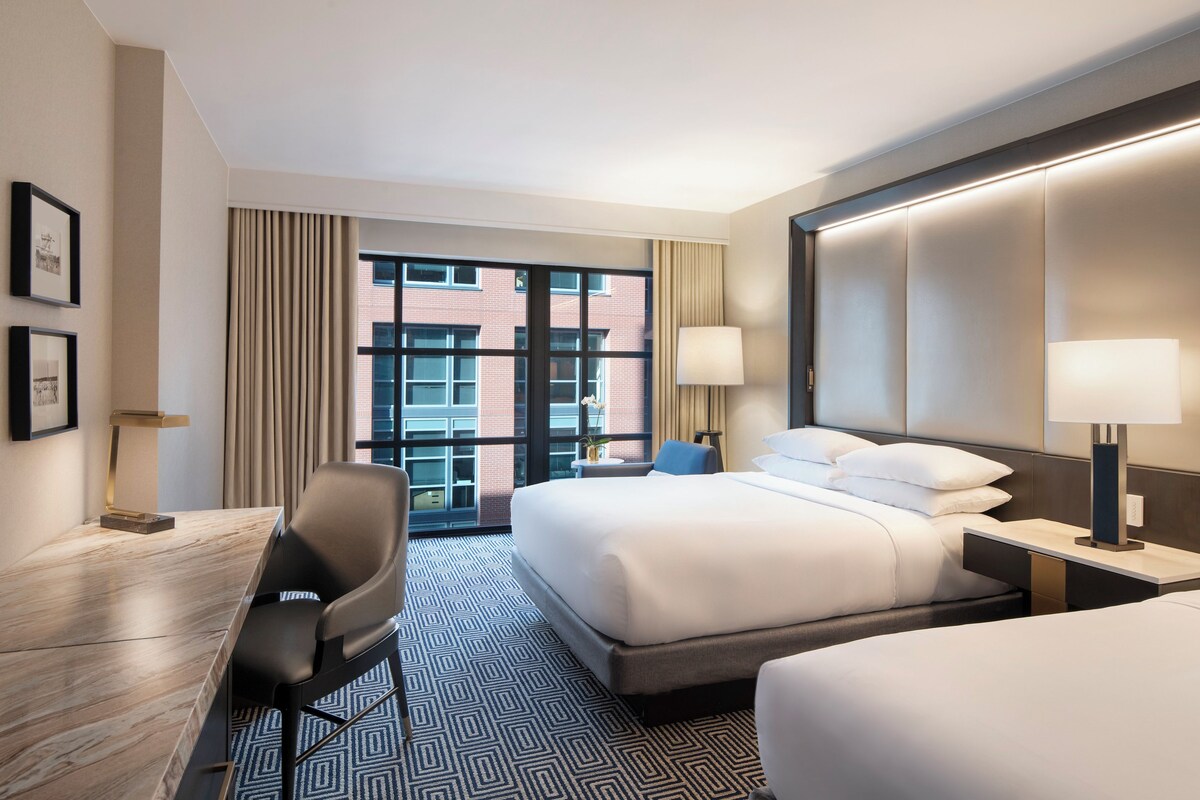 A hotel room with a bed, two bedside tables, a desk, a chair, and a view of the outside.