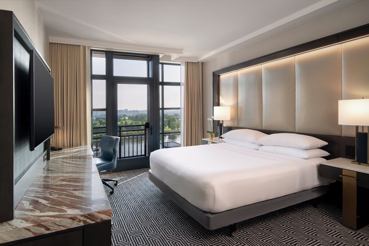 A hotel room with a large bed, a desk, a TV, and a view of a lake.