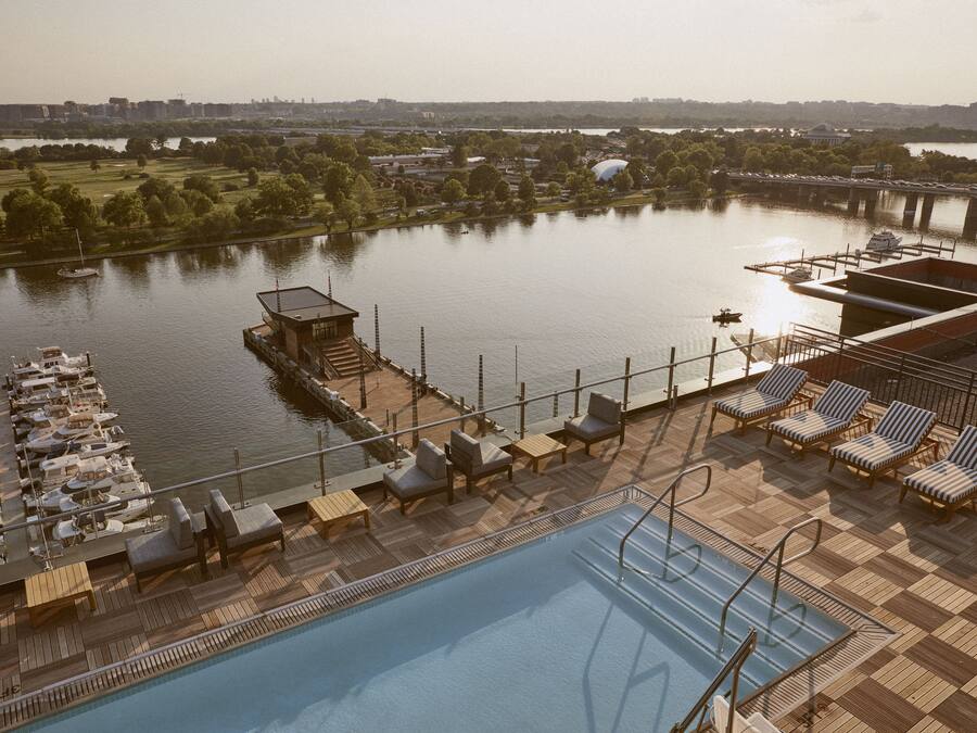 A rooftop pool area with lounge chairs and a swimming pool, overlooking a river with a bridge and greenery in the background.