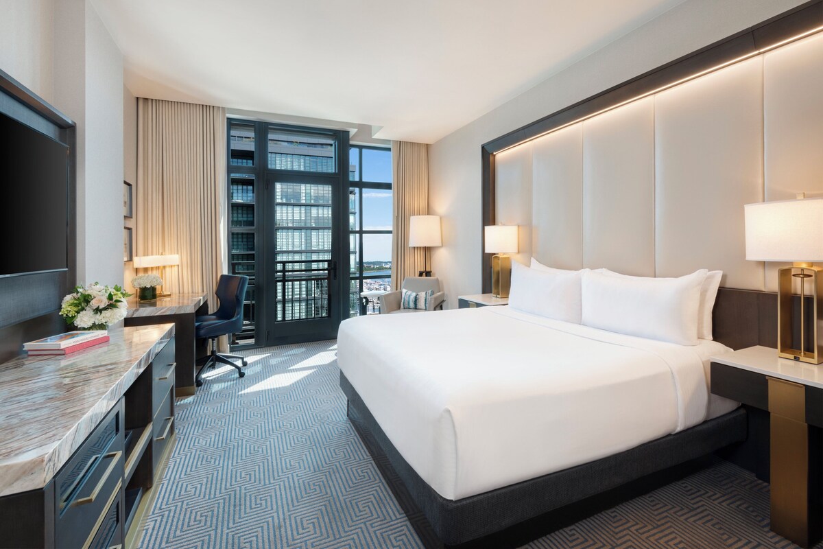 A hotel room with a large bed, a desk with a chair, a TV, and a view of the city through a large window.
