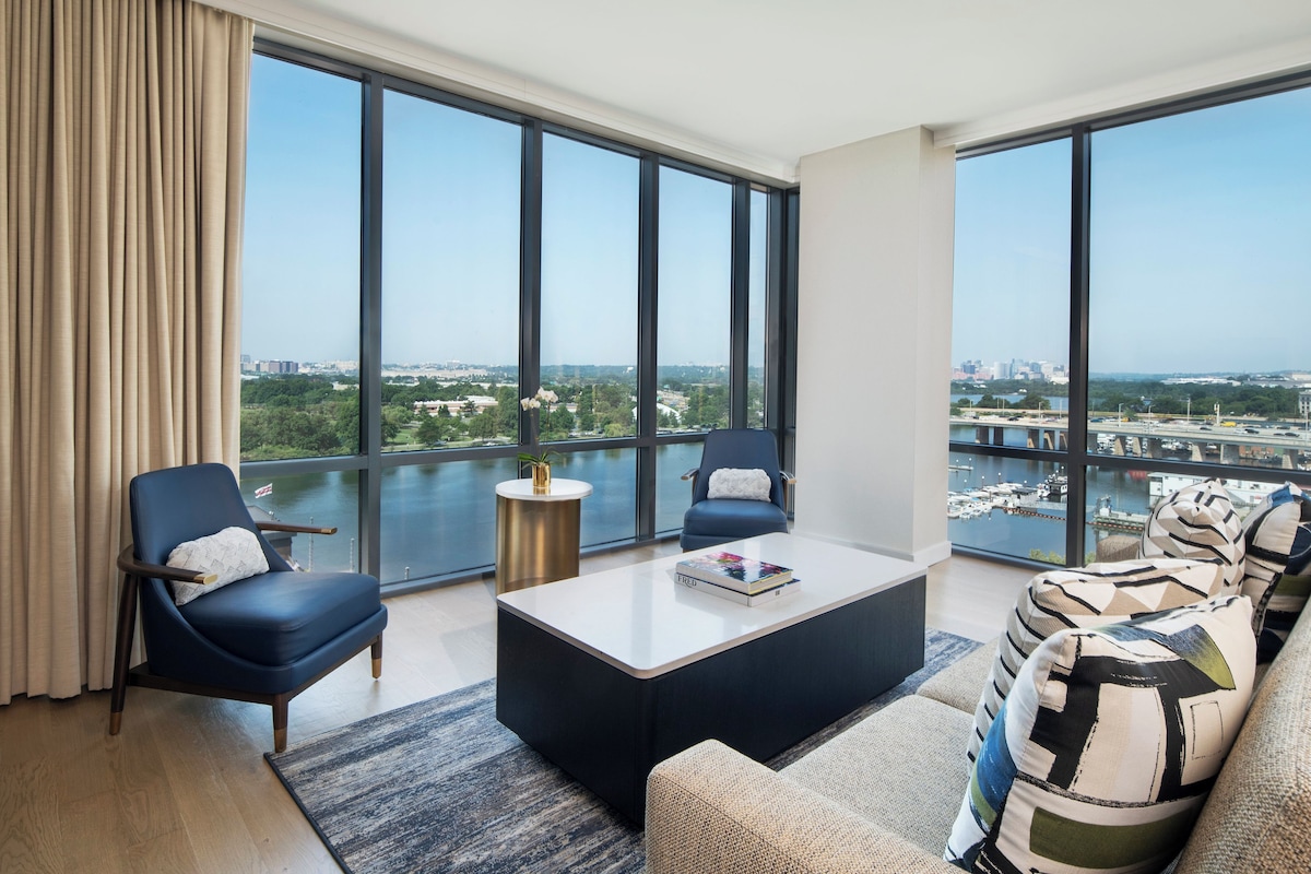 A modern living room with a large window, a sofa, two armchairs, a coffee table, and a view of a cityscape and river.
