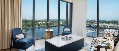 Una sala de estar moderna con un ventanal grande, un sofá, dos sillones, una mesa de centro y vistas a un paisaje urbano y un río.