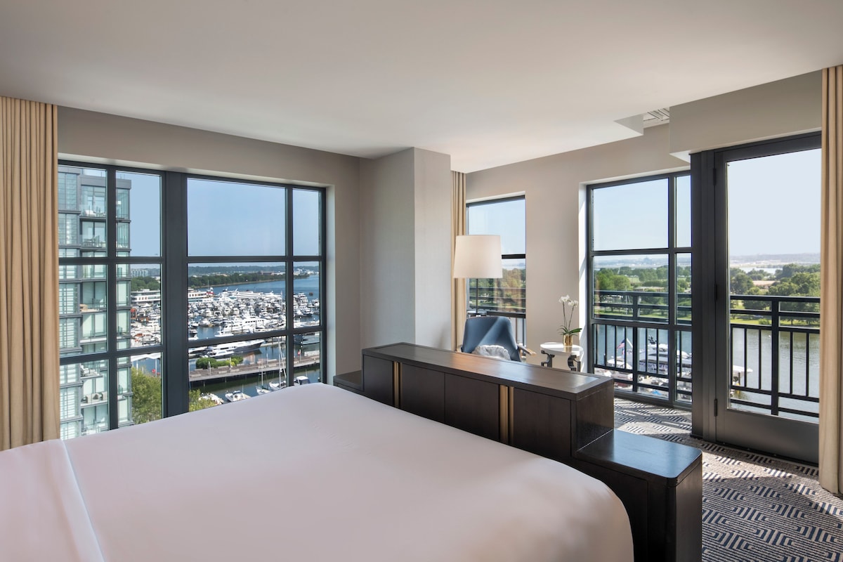 A hotel room with a large bed, a desk, and a view of a marina and cityscape through large windows.