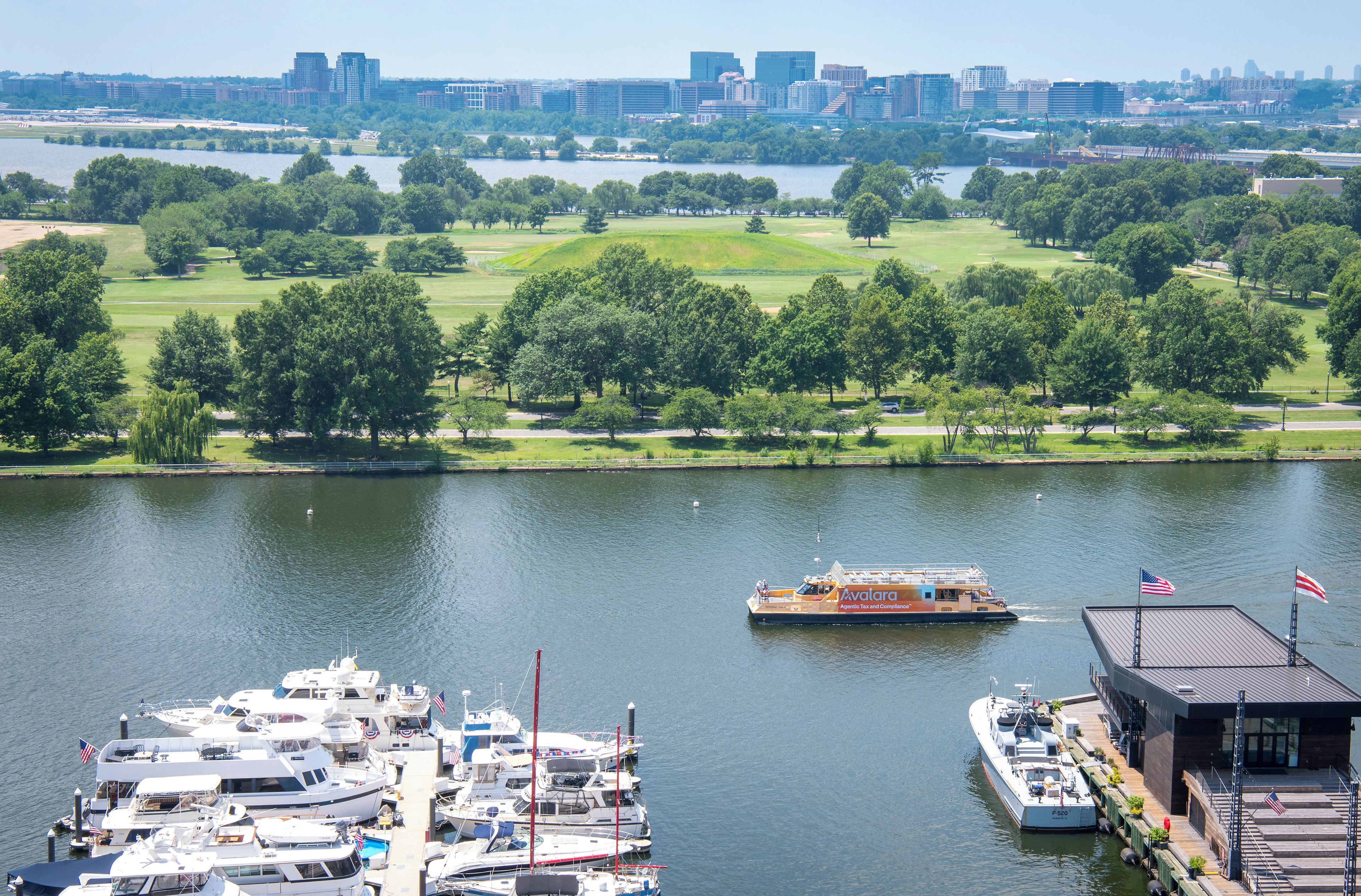 A marina with boats docked, a golf course in the background, and a city skyline across the water.