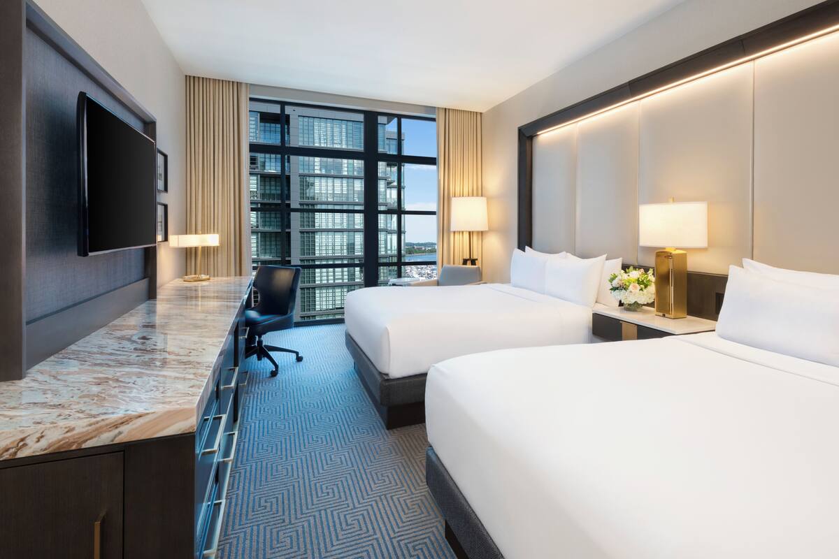 A hotel room with two beds, a desk with a flat-screen TV, a chair, and a large window with a city view.