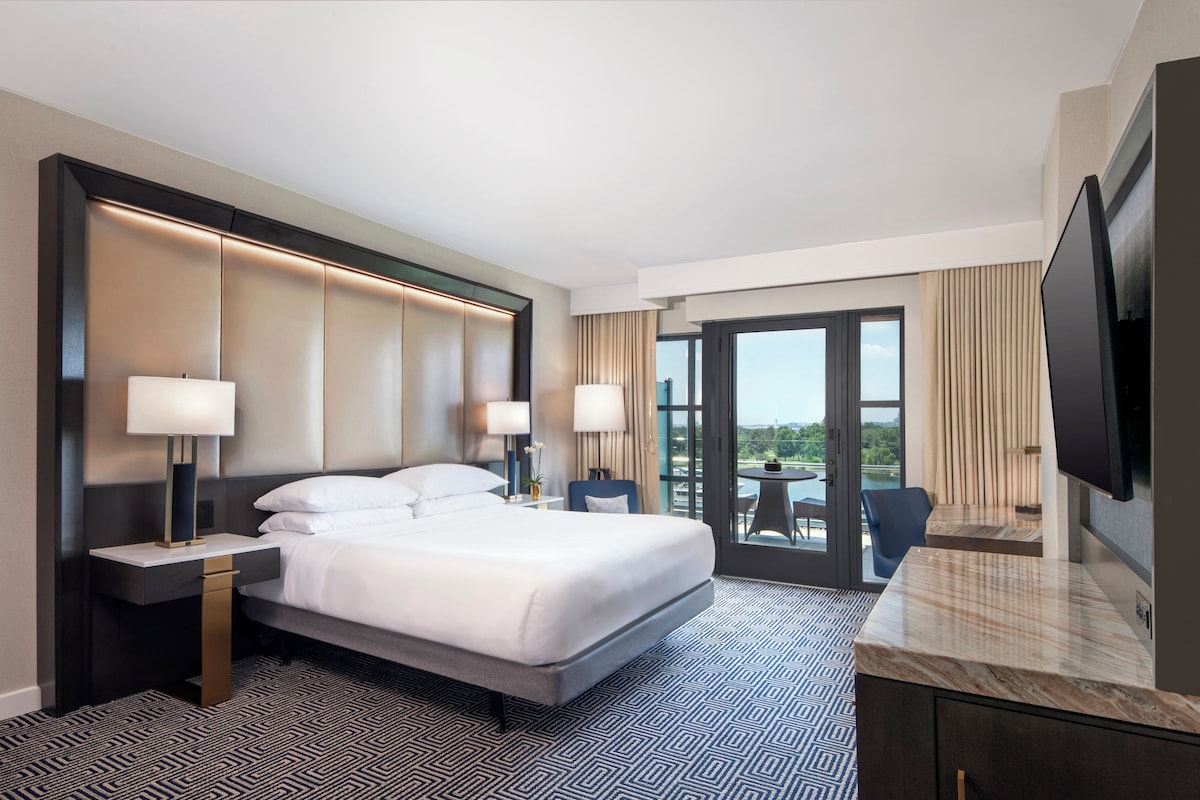 A hotel room with a large bed, two bedside tables with lamps, a TV, and a view of a body of water through the window.
