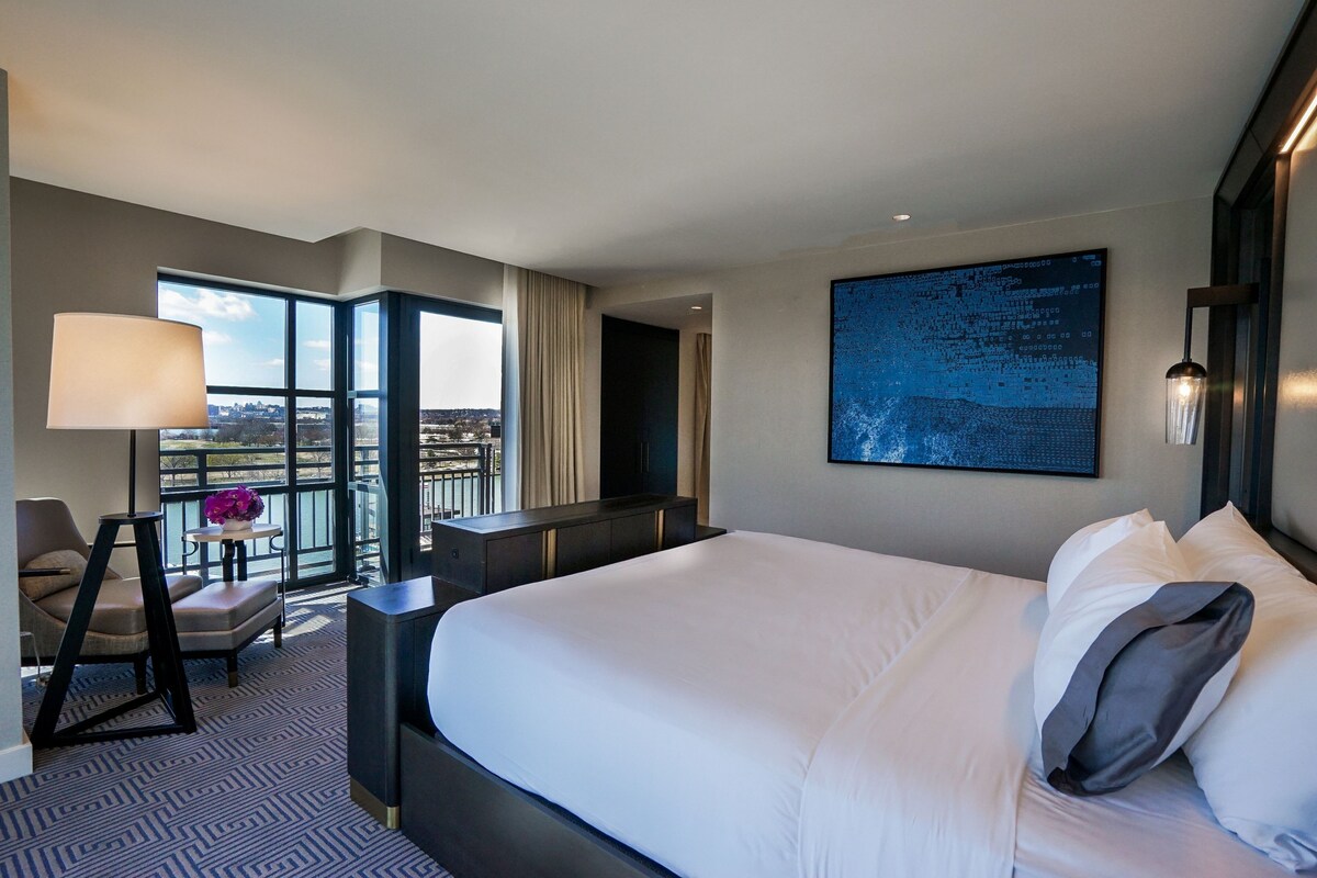 A hotel room with a large bed, a desk, a chair, a view of the city, and a large abstract painting on the wall.