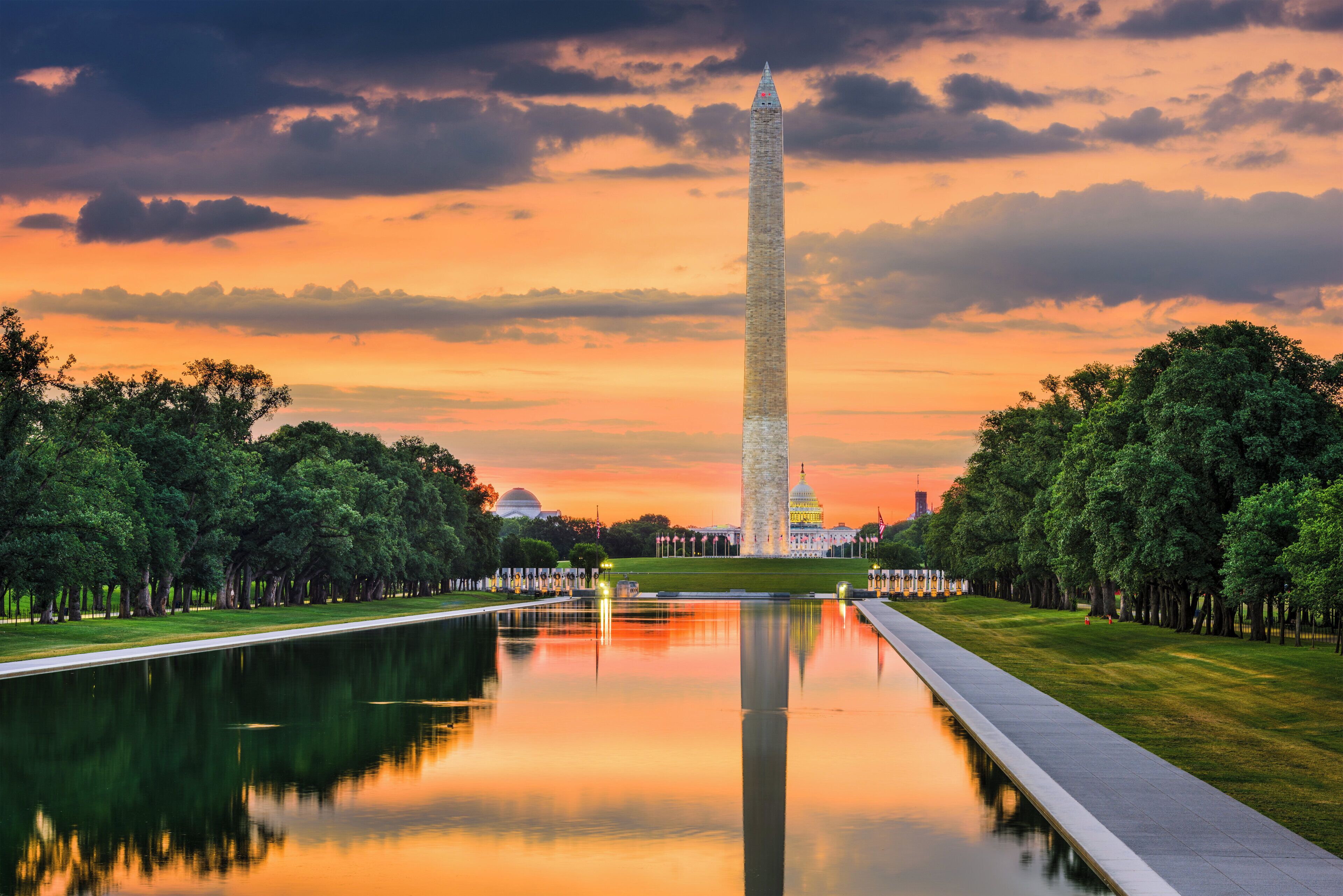 Le Washington Monument se reflète dans une étendue d’eau calme, entouré d’arbres et sous un ciel dégagé.