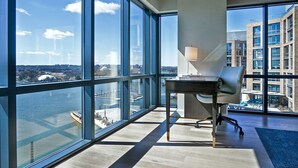 Un espacio de oficina moderno con un amplio ventanal que da a un río y al paisaje urbano, un escritorio con lámpara y vistas de edificios contiguos.