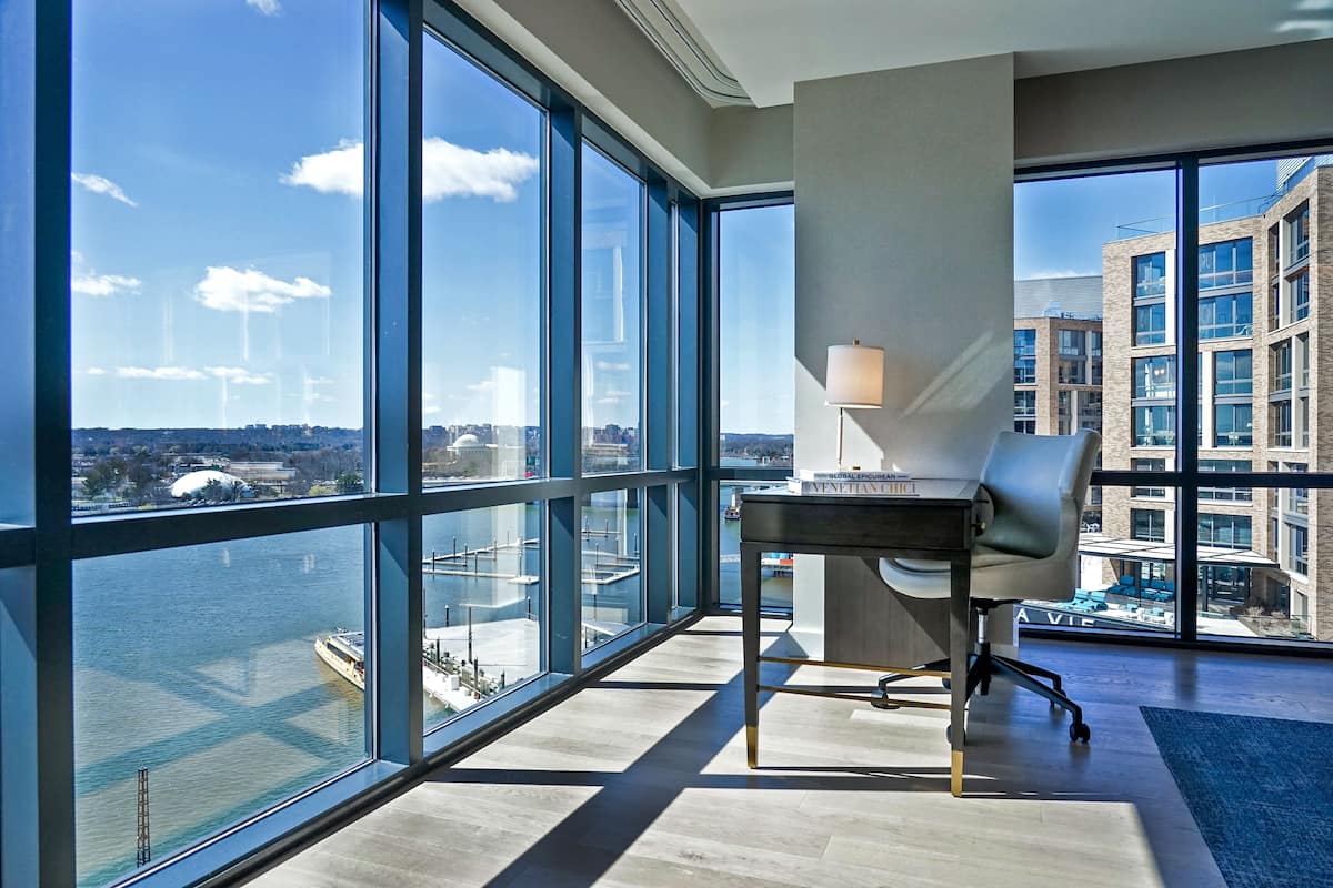 A modern office space with a large window overlooking a river and cityscape, a desk with a lamp, and a view of adjacent buildings.