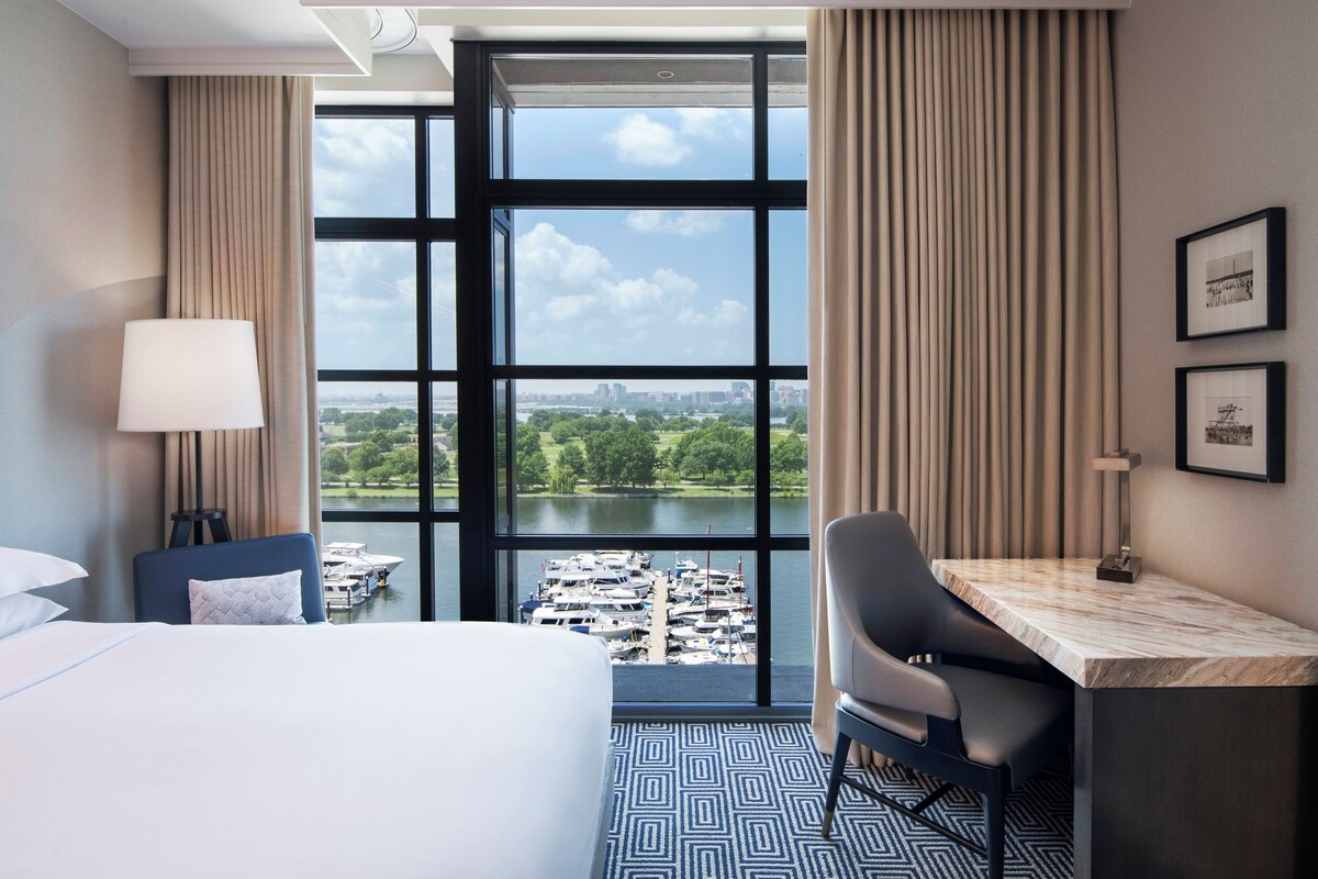 A hotel room with a large window, a bed, a desk, a chair, and a view of a lake and cityscape.