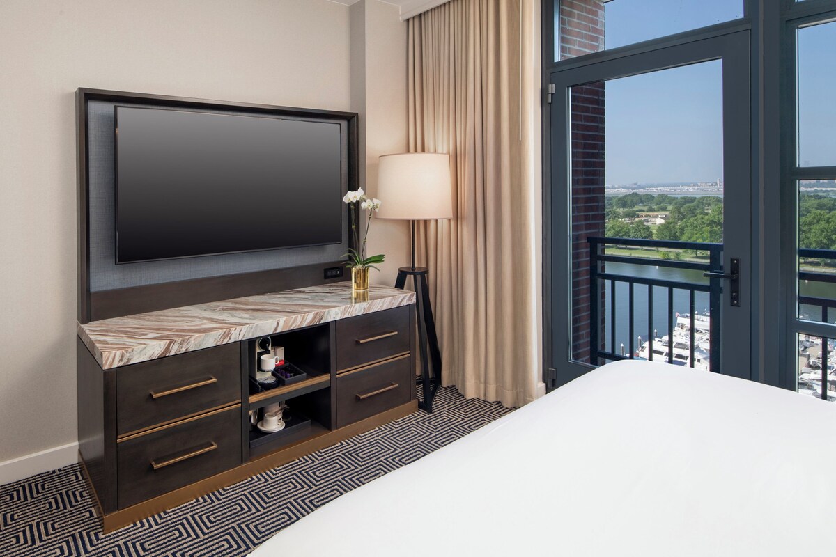 A hotel room with a large flat-screen TV mounted on the wall, a bedside table with a lamp, a vase with flowers, and a view of a cityscape through a glass door.