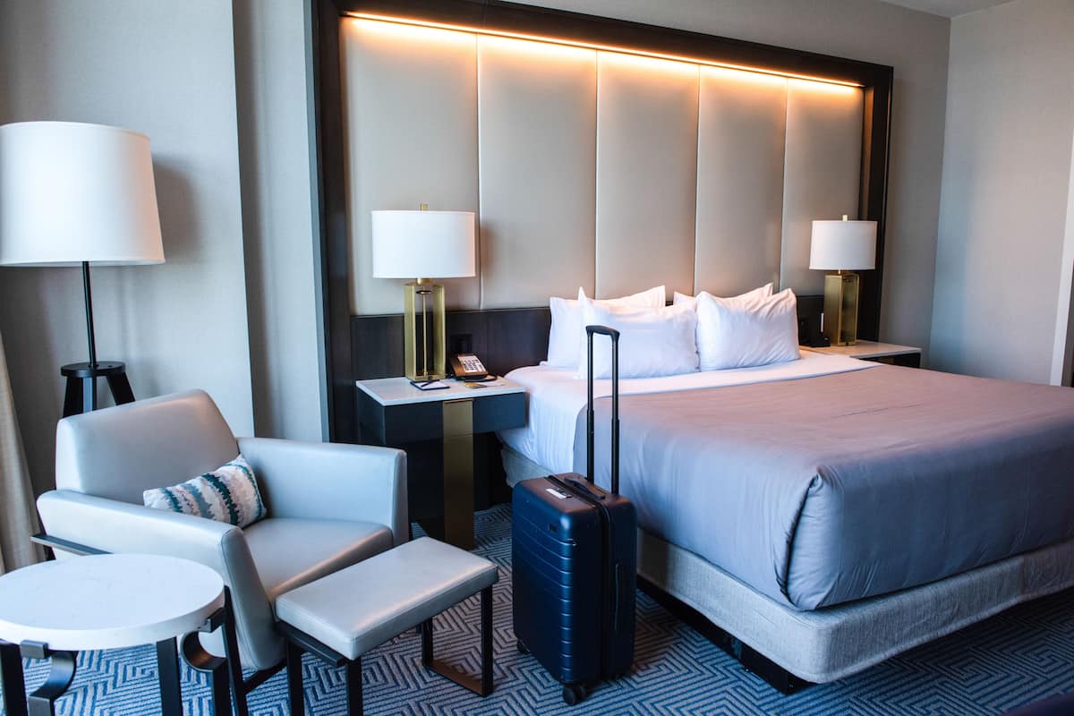 A hotel room with a bed, a desk, a chair, a table, and a suitcase.