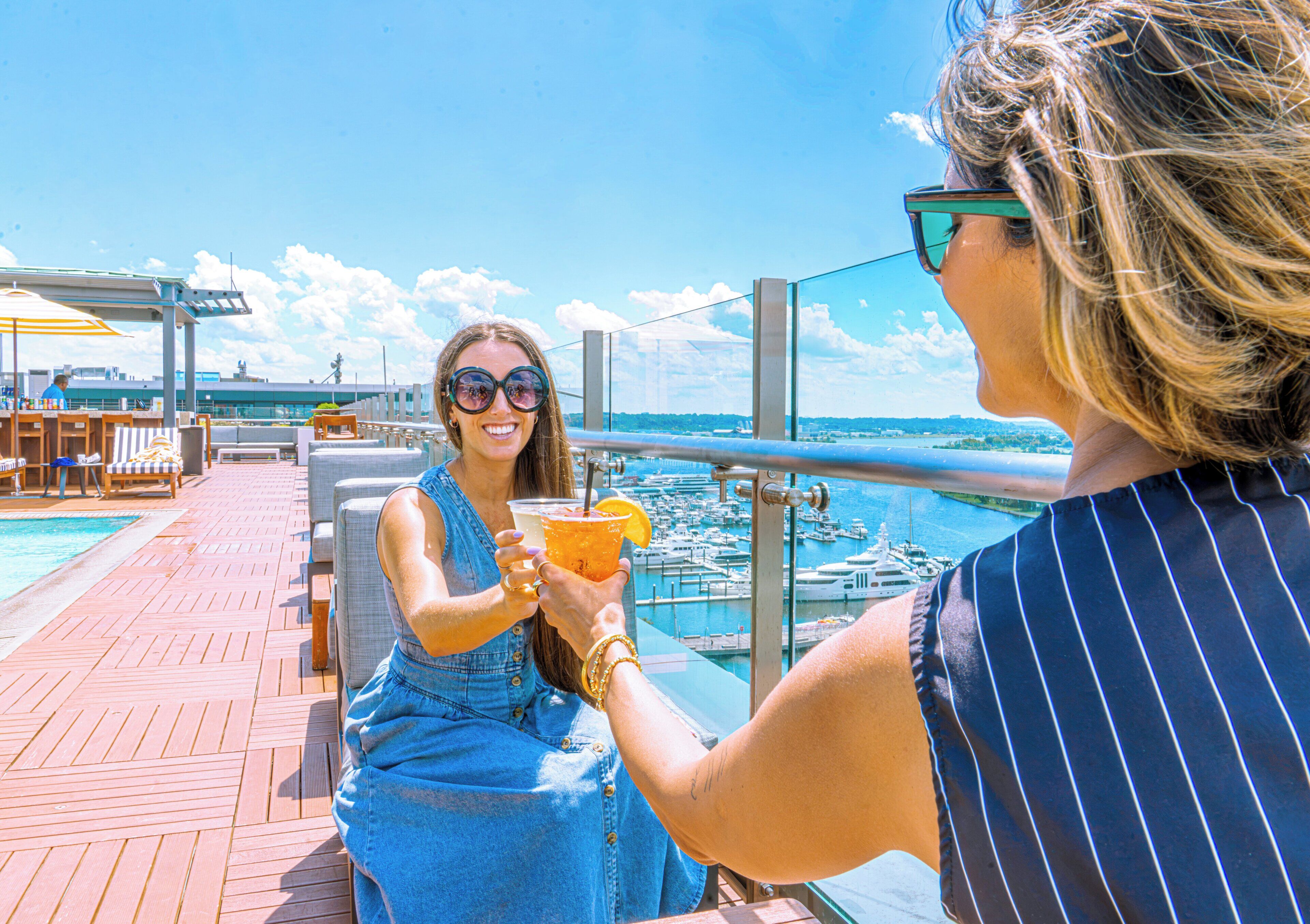 Une personne tenant un verre contenant une tranche de fruit, assise sur une terrasse avec vue sur un port de plaisance et des bateaux.