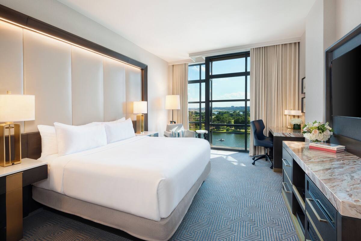 A hotel room with a large bed, two bedside tables with lamps, a desk with a chair, a TV, and a view of the outdoors.