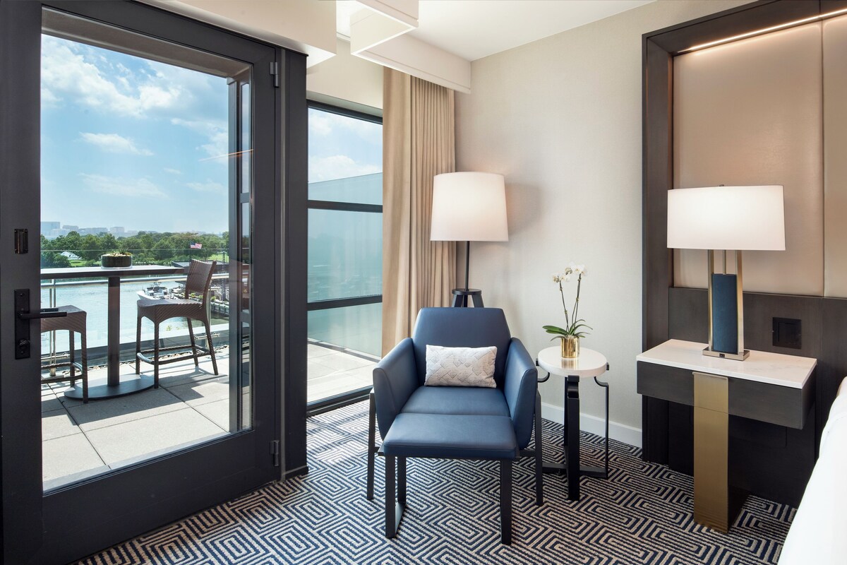 A hotel room with a view of a marina, a seating area with a table and chairs, a blue armchair, a small round table with a lamp, and a bed with a bedside table.