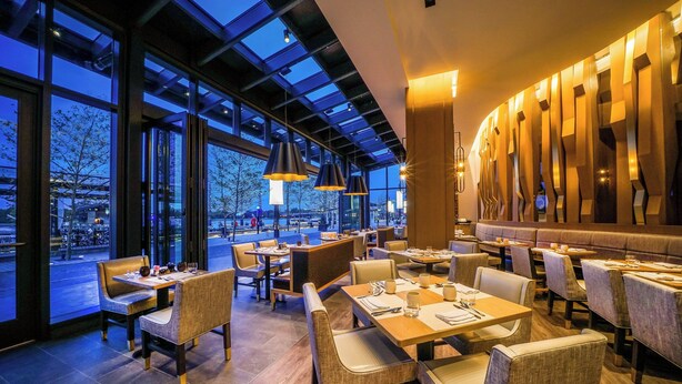 Un restaurante moderno con amplios ventanales, mesas de madera y sillas acolchadas.