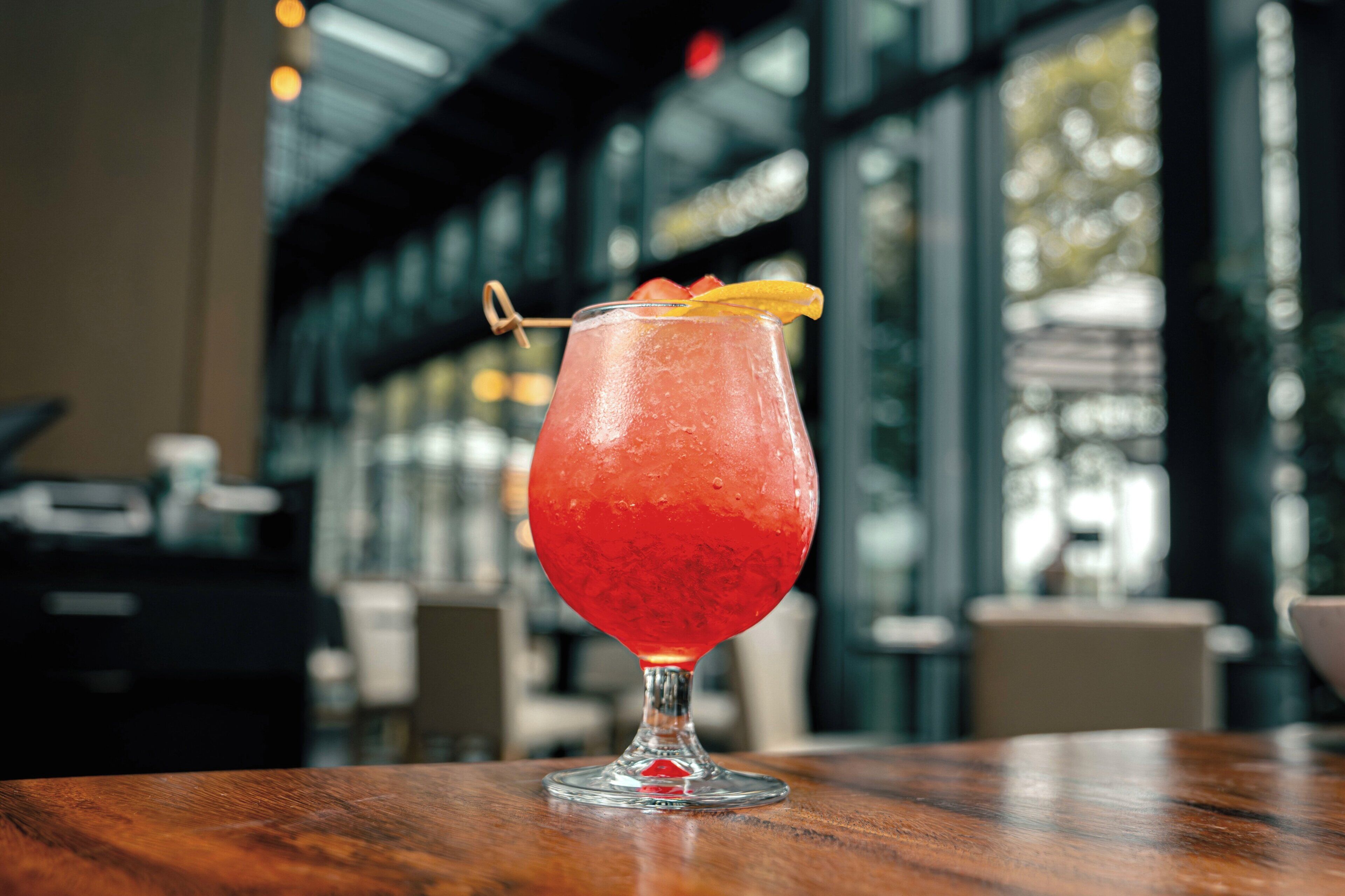 A cocktail in a glass with a garnish on a wooden table in a modern interior with large windows.