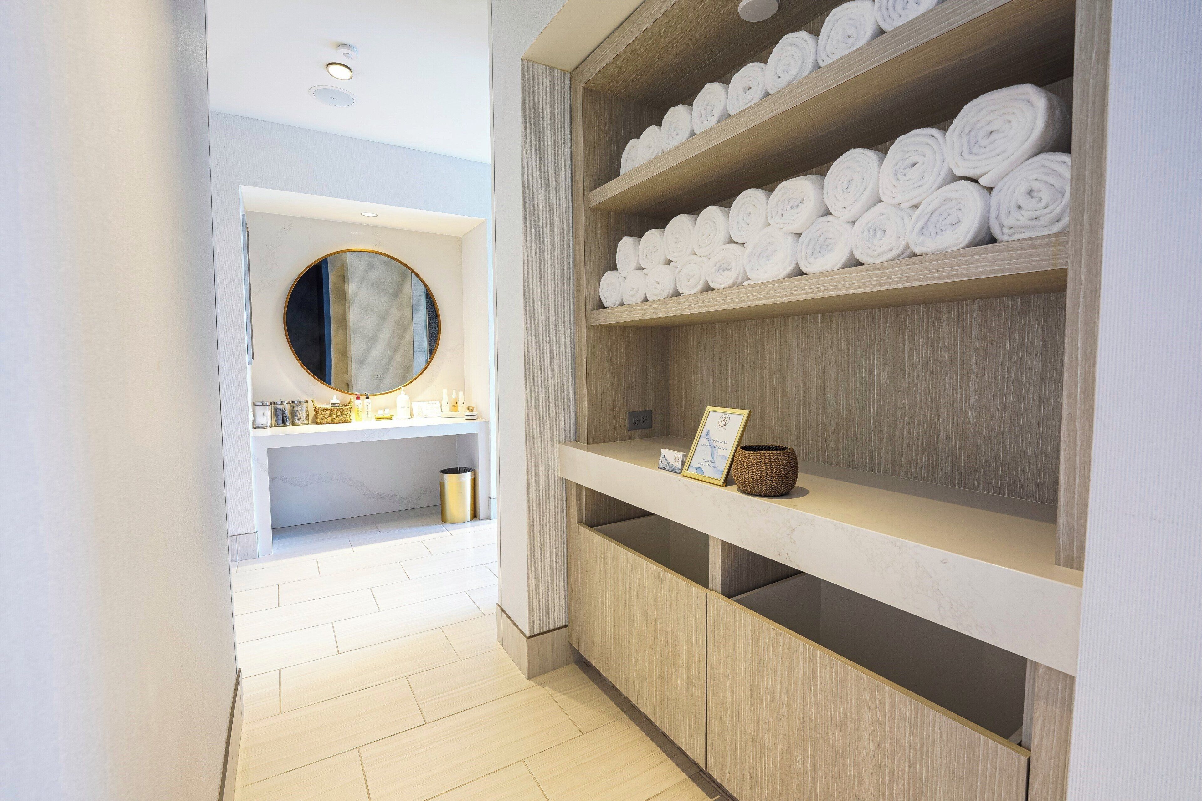 A hotel bathroom with wooden shelves holding rolled towels, a round mirror, and a small shelf with toiletries.