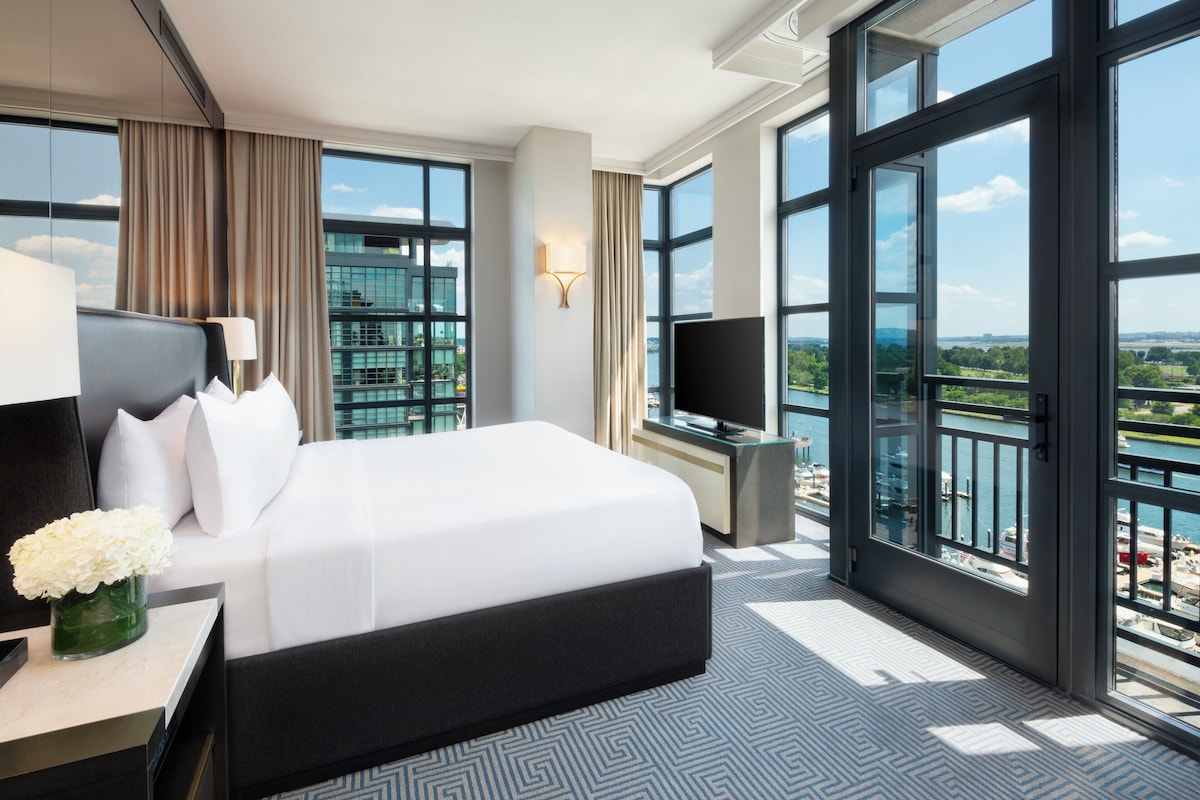 A hotel room with a large bed, a desk with a lamp, a TV, and a view of a body of water through large windows.