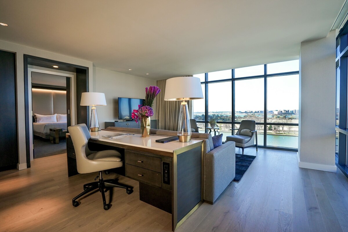 A modern hotel room with a desk, chair, sofa, and large windows offering a city view.